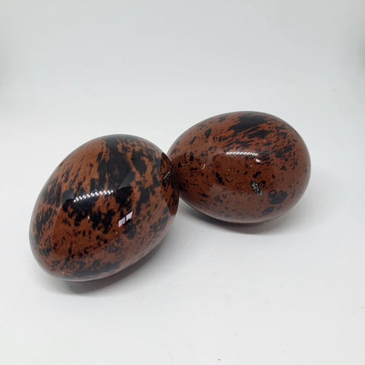 Two mahogany obsidian eggs with a reddish-brown color and black patterns, placed on a white surface.